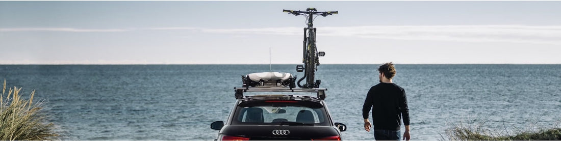 A person stands next to a black car with a Thule bicycle mount on its roof near the coast. The car faces the ocean, and the person is looking at the water.