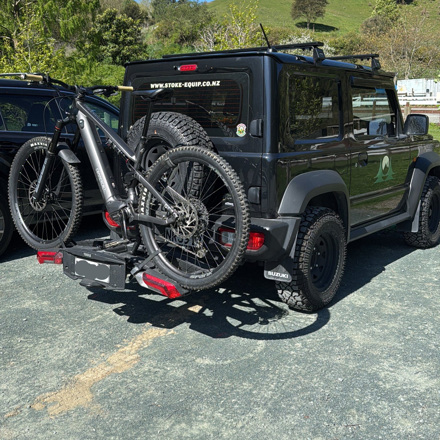 Black Suzuki Jimny with a bike rack and mountain bike at codgers mtb trails in nelson, with a clear blue sky and greenery.