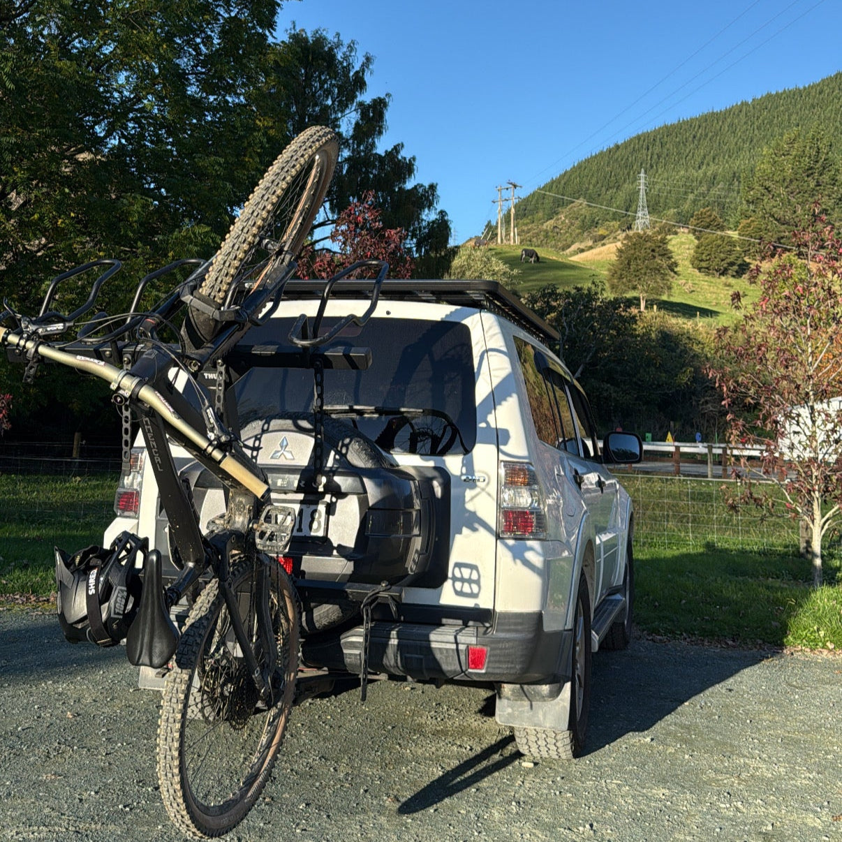 White SUV with a bike rack carrying two bicycles on a road with trees and mountains in the background.