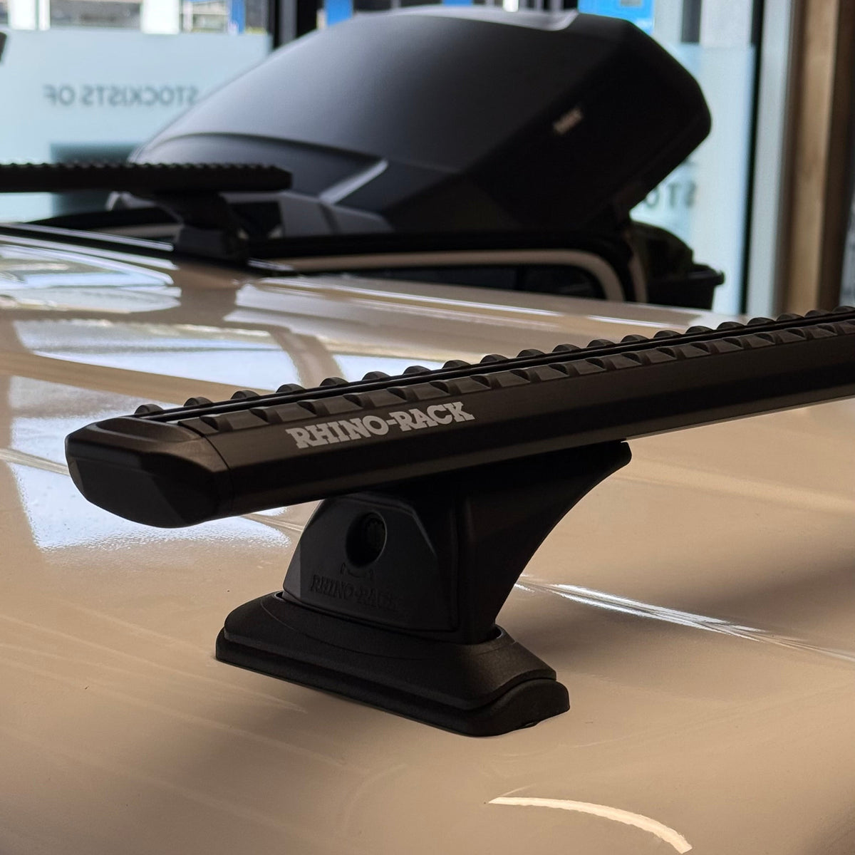 A black Rhino-Back roof rack bar mounted on the white roof of a vehicle, with a black roof box visible in the background inside a showroom.
