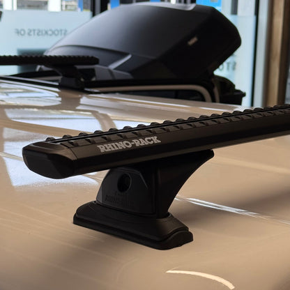 A black Rhino-Back roof rack bar mounted on the white roof of a vehicle, with a black roof box visible in the background inside a showroom.