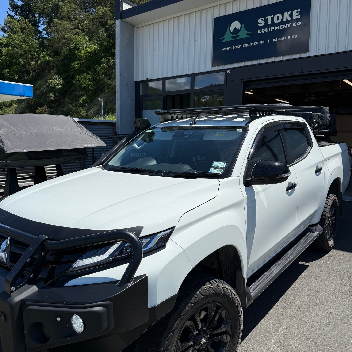 White Mitsubishi Triton, fitted with a Rhino-Rack Pioneer Platform Roof Tray and RMTB1 Backbone mounting system, parked outside Stoke Equipment Co, 74b St Vincent Street Nelson