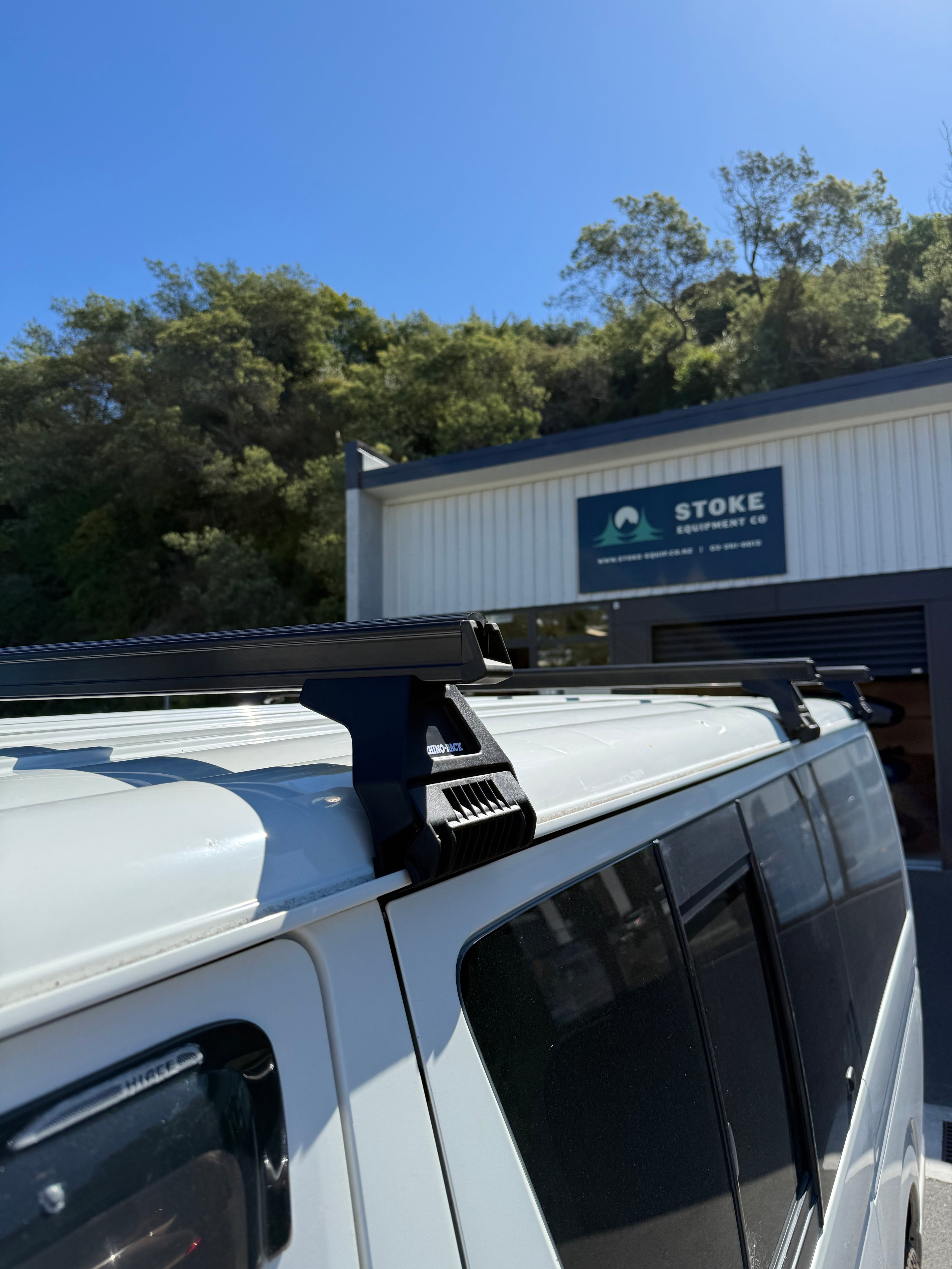White Suzuki Jimny with Rhino-Rack HD ROof Racks, parked in front of Stoke Equipment Co building with a blue sky and trees in the background