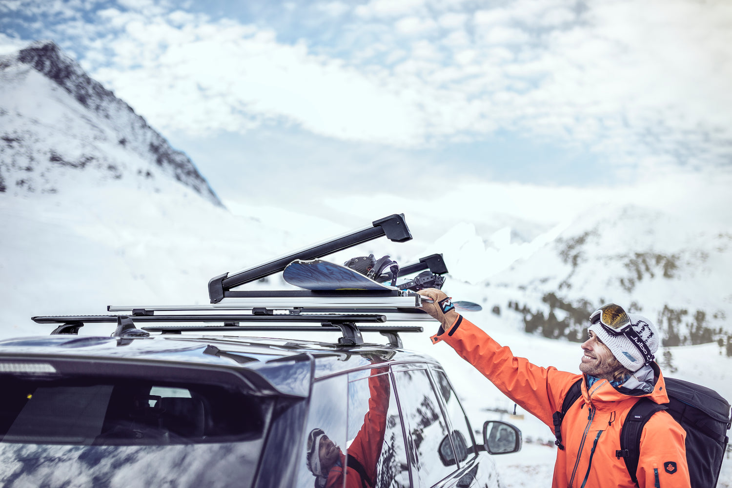 Image of a man loading a snowboard onto his thule snowpack ski and snowboard rack. parked at the base of a ski filed 