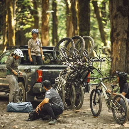 Mountain bikers with several bicycles on a carrier rack and in the bed of a pickup truck in a forest setting. One person is crouching near a dog, while others stand or sit with the bikes.