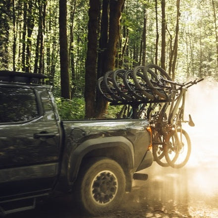 A black pickup truck on a dirt forest road carrying multiple bicycles on a rack mounted behind the bed, with bright sunlight and dust in a wooded outdoor setting.