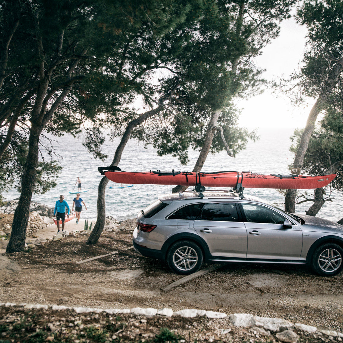Thule Kayak Racks mounted on an Audi Allroad parked next to the beach, with people carrying an orange kayak to load onto the vehicle