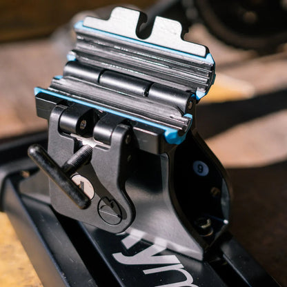 Close-up of a black metal mounting bracket with blue accents and a thumbs-toggle screw, attached to a black rail in an outdoor setting.